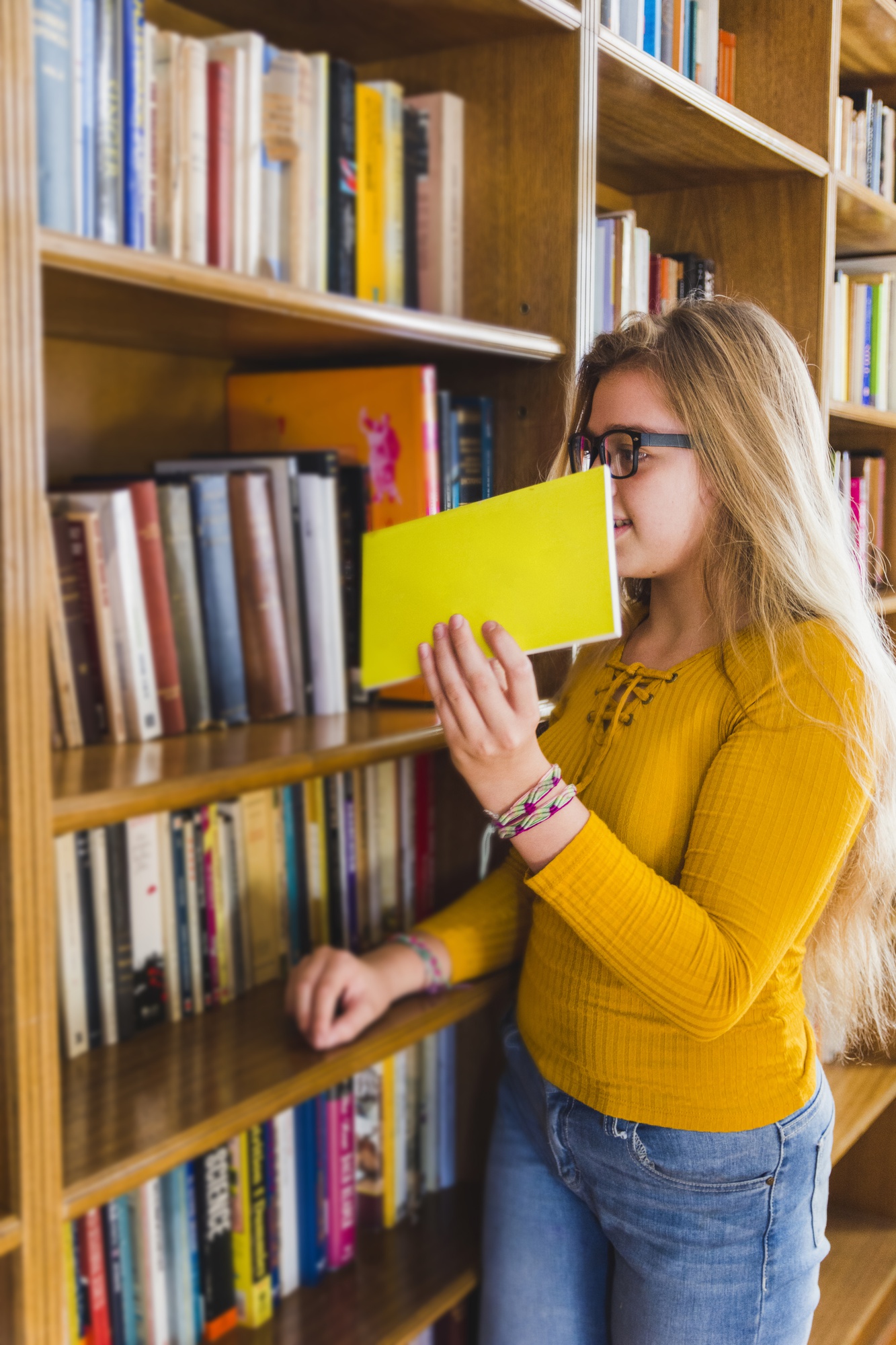 Jovem em estante de biblioteca segurando um livro de capa amarela lisa, cercada de livros coloridos nas prateleiras.
