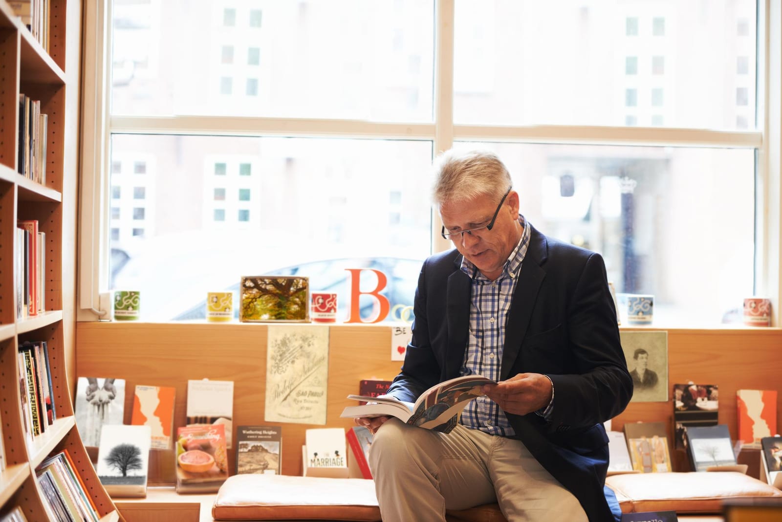 Homem sênior sentado em banco de livraria lendo um livro ao lado de estantes com capas coloridas.