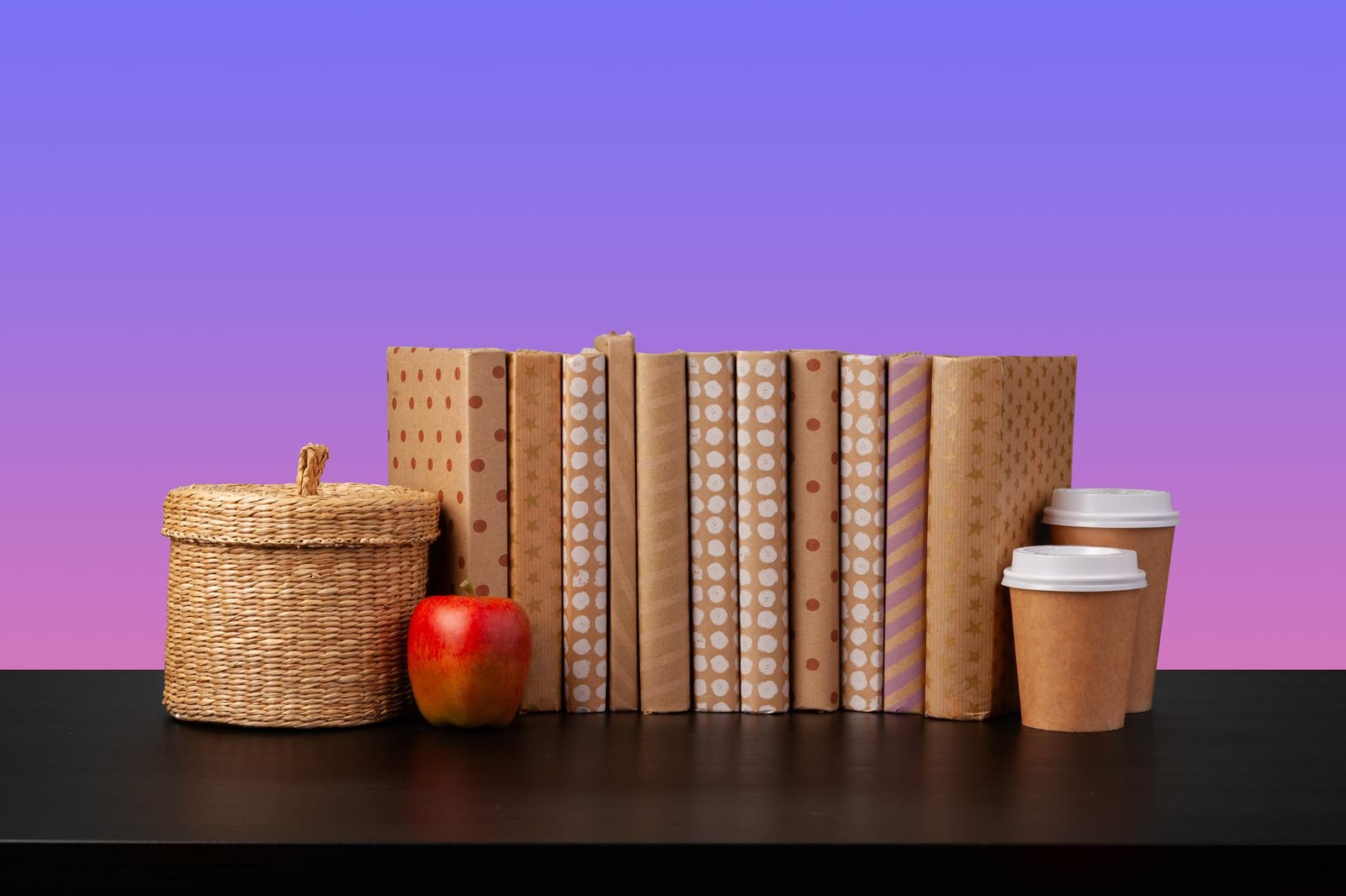 Livros embrulhados em papel kraft padronizado ao lado de uma cesta de palha, uma maçã e copos de café para viagem.
