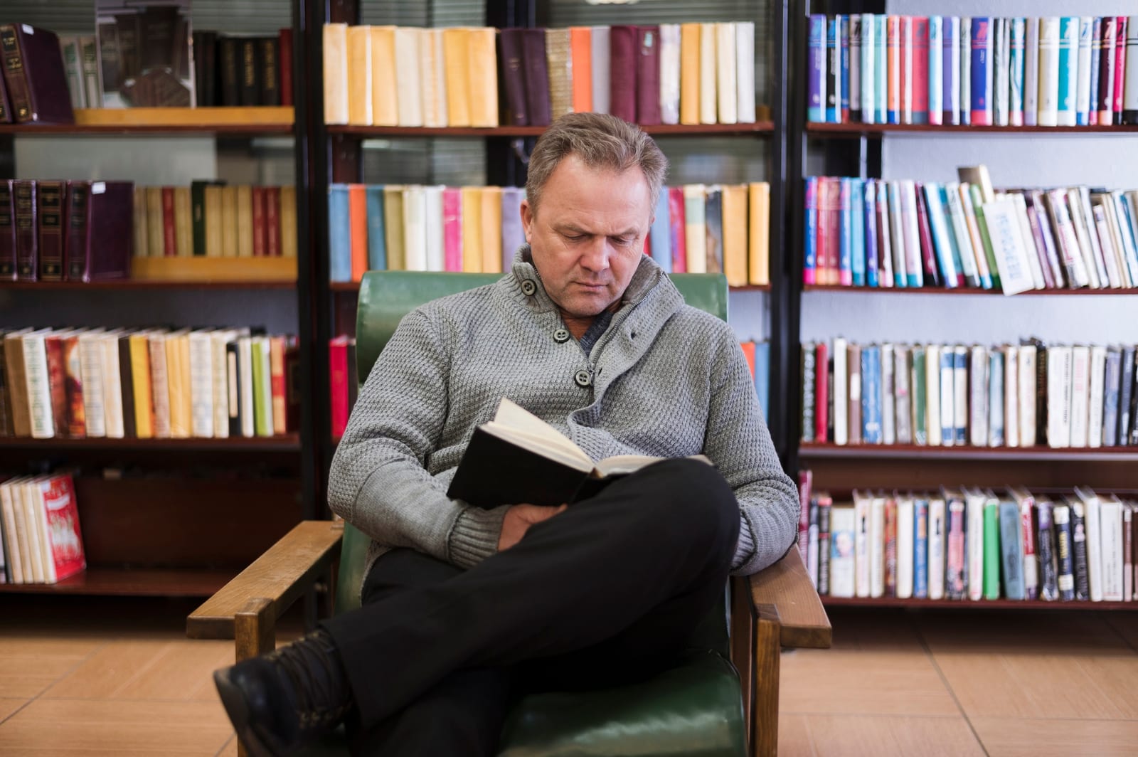Homem lendo um livro em poltrona de couro verde, cercado por estantes cheias de livros coloridos.