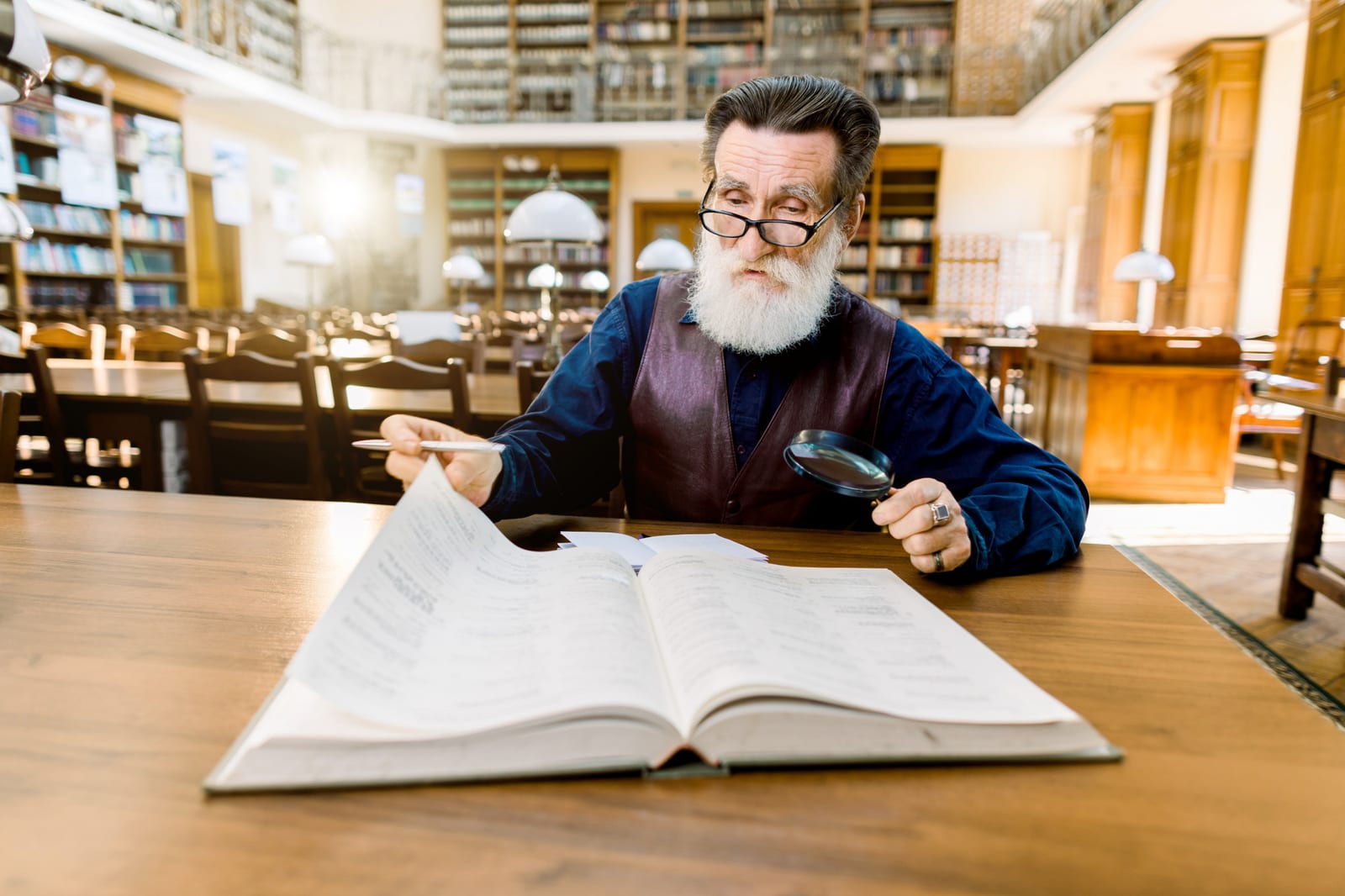 Homem mais velho em biblioteca antiga, lendo um livro grande sobre a mesa com o auxílio de uma lupa.